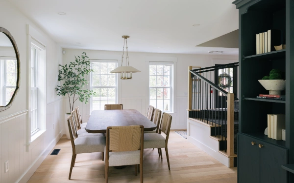 Wide library and dining view featuring dark built-in shelving, stair railing detail, long wood table, and natural light from multiple windows